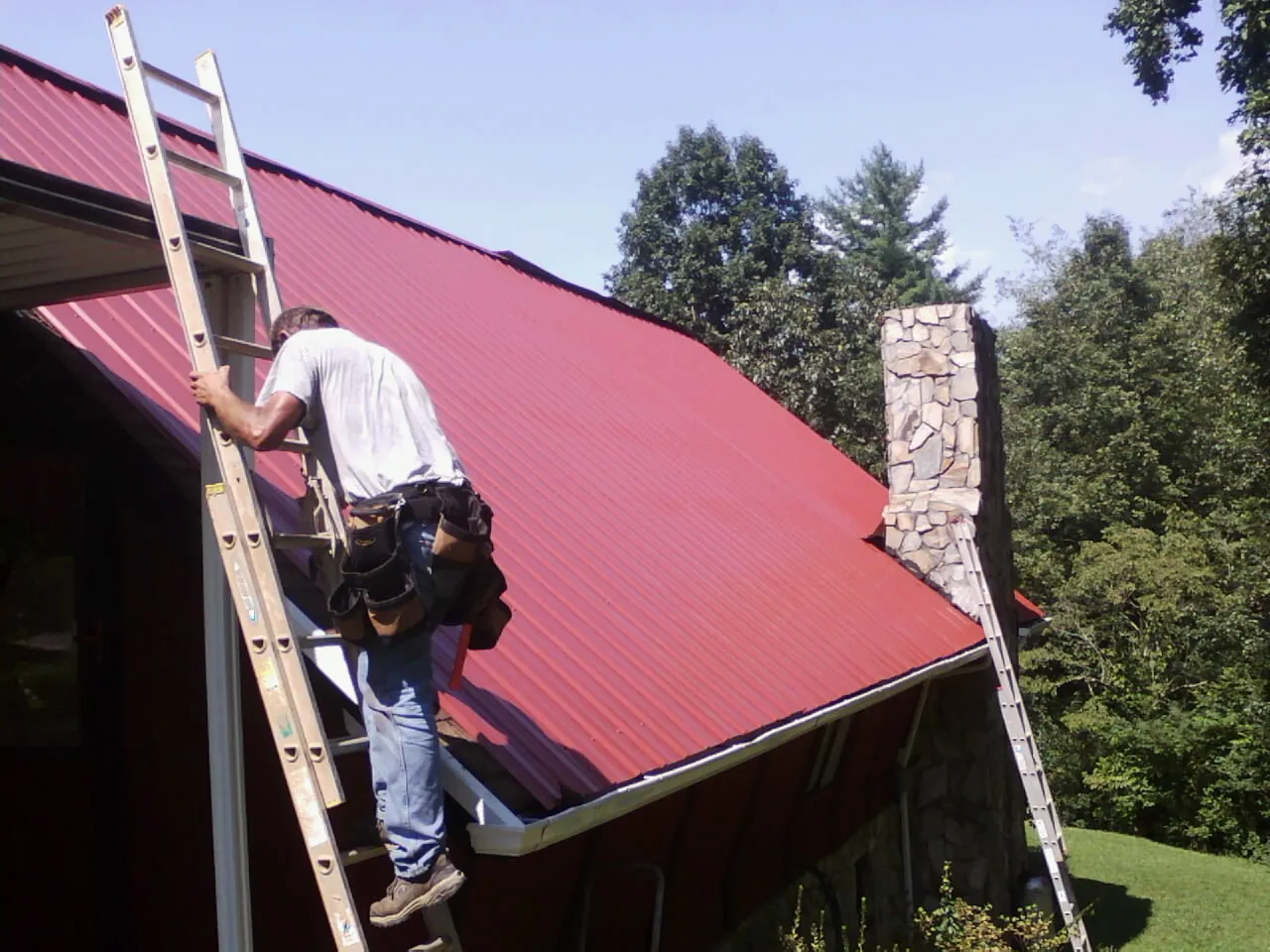 Roofer on a ladder beside a red metal roof and stone chimney — Soffit & Fascia Repair by Roofing Brothers in Newberry